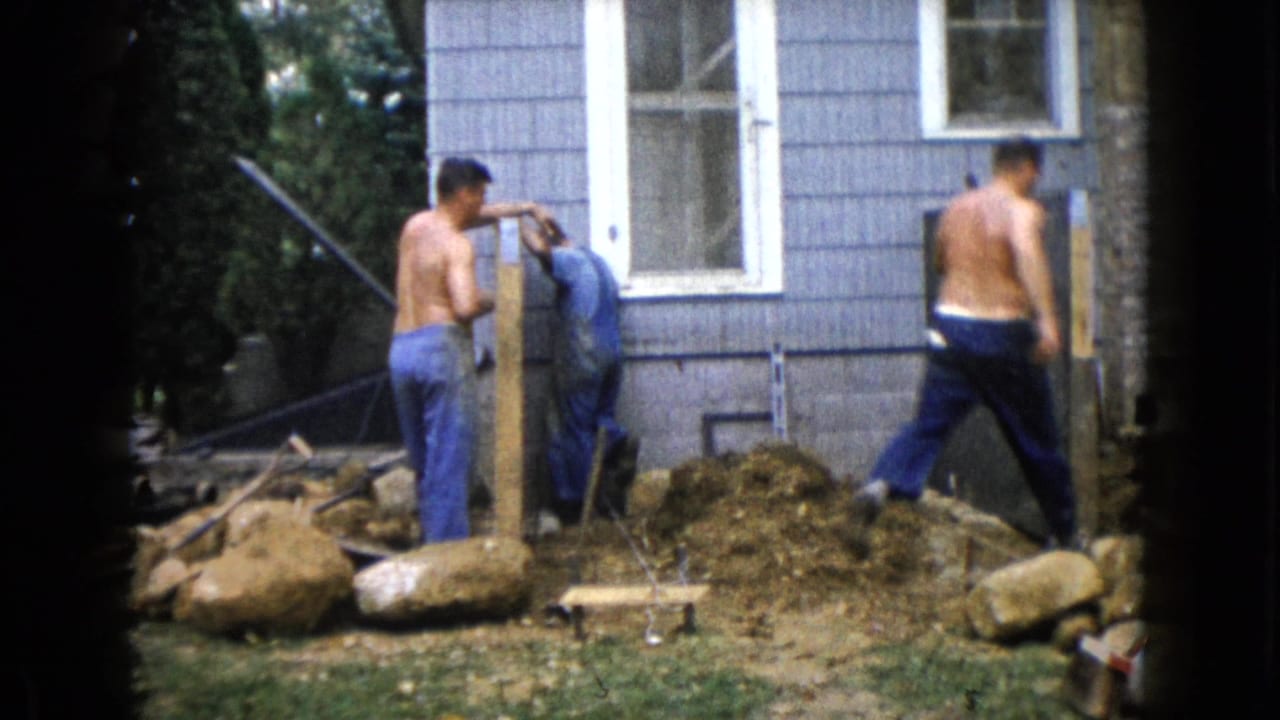 Two men work beside a wooden post in a backyard next to a blue-sided house.