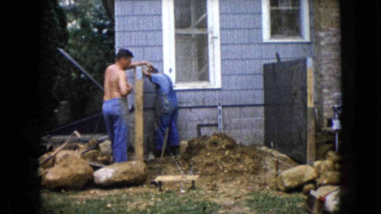 1960 Wauconda frame with a shirtless man and a man in blue overalls working near a wooden post outside a blue house.