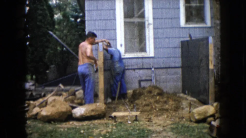 1960 Wauconda backyard construction frame showing two men at a wooden post, with a blue house wall, rocks, soil, and tools nearby.