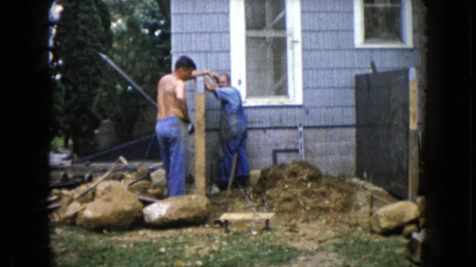 1960 Wauconda home-movie frame of two men beside a wooden post, with one turning toward the camera near a blue-sided house.
