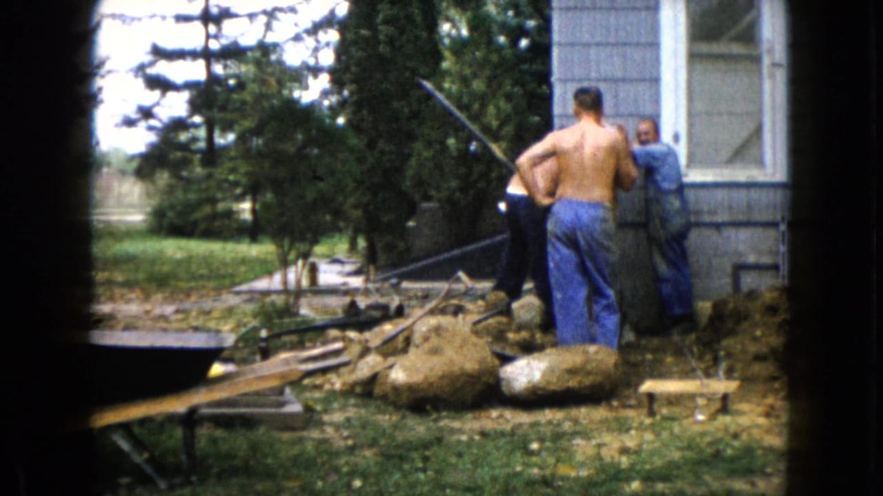 Two shirtless men and a man in a blue shirt stand near rocks, dirt, and tools by a house.