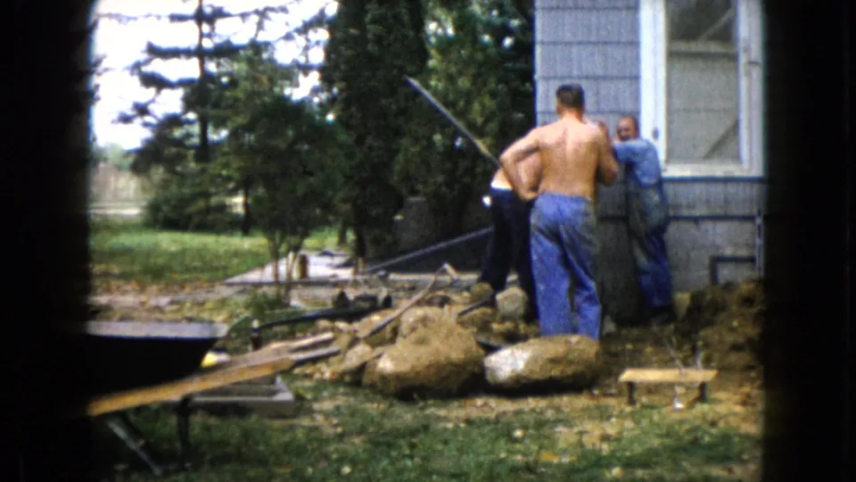Three adult men continue outdoor work beside a gray house in 1960 Wauconda, with rocks, posts, and scattered tools in view.