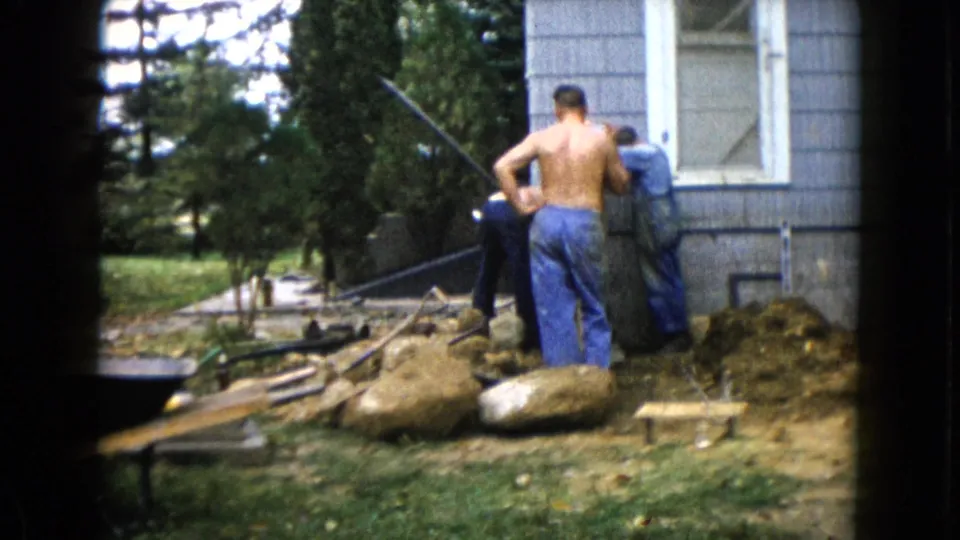 A 1960 home-movie frame shows men working among rocks, boards, and dirt beside a gray-sided house in Wauconda.