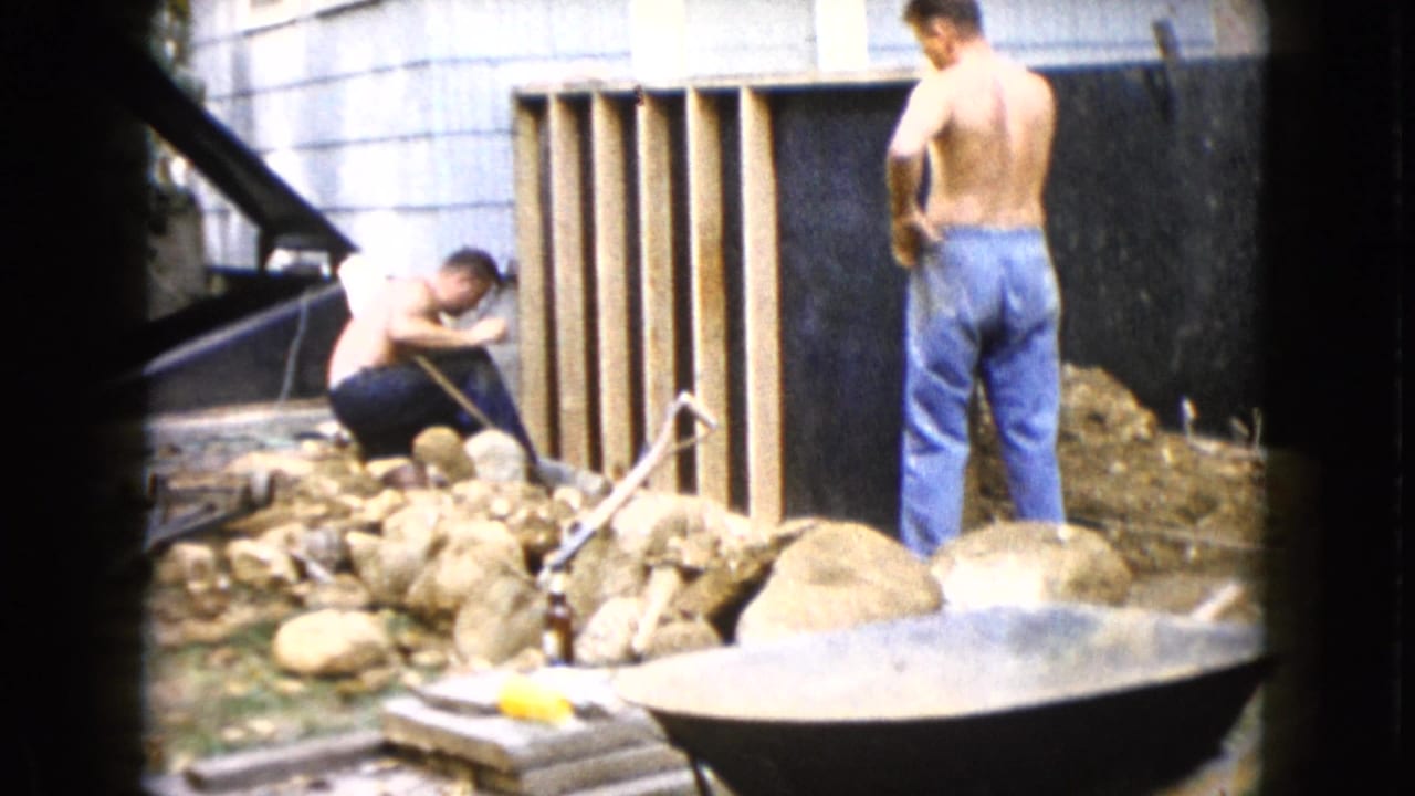 In 1960 Wauconda, Illinois home-movie footage, two shirtless men work beside a wooden frame in a rocky backyard with a wheelbarrow nearby.