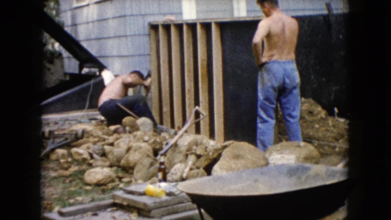 A shirtless man crouches at the base of a wooden frame while another stands nearby in 1960 Wauconda backyard home-movie footage.