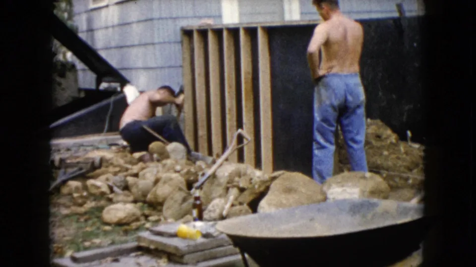 A shirtless man crouches at the base of a wooden frame while another stands nearby in 1960 Wauconda backyard home-movie footage.
