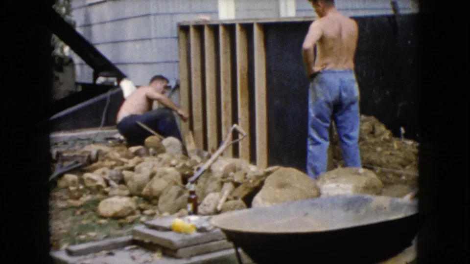 The 1960 Wauconda home movie shows a backyard work area with a crouching man, standing man, wood framing, rocks, and exposed soil.