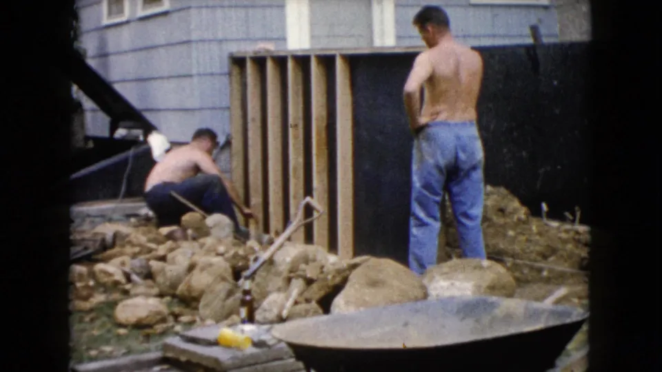 A shirtless man reaches down near the yard frame while another stands nearby in 1960 Wauconda, Illinois backyard work footage.
