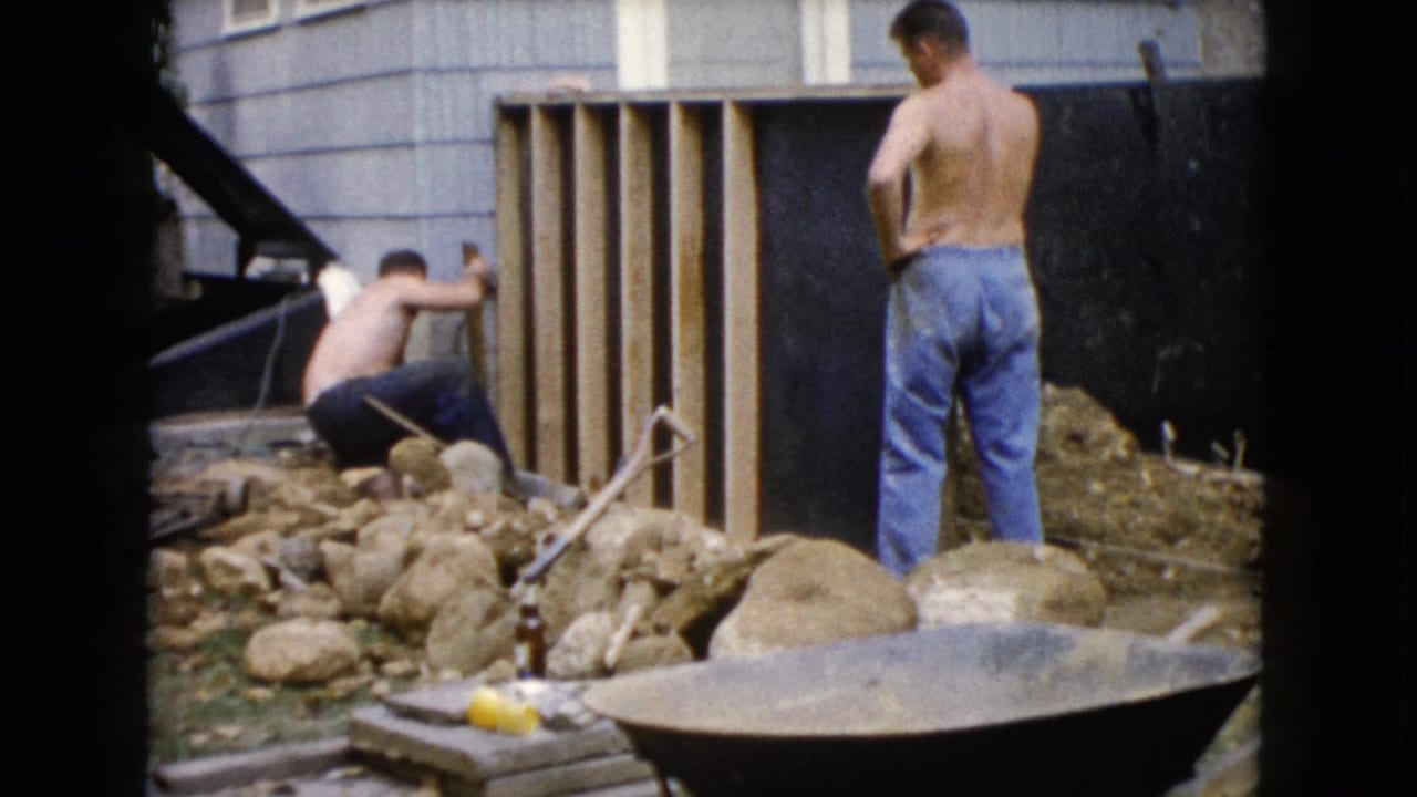 Two shirtless men work beside a wooden frame in a rocky residential yard.