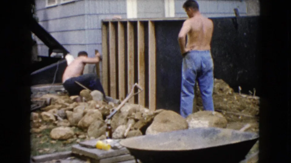 In Wauconda, Illinois, 1960 home-movie footage, two men work by a wooden structure near house siding, rocks, dirt, and a wheelbarrow.