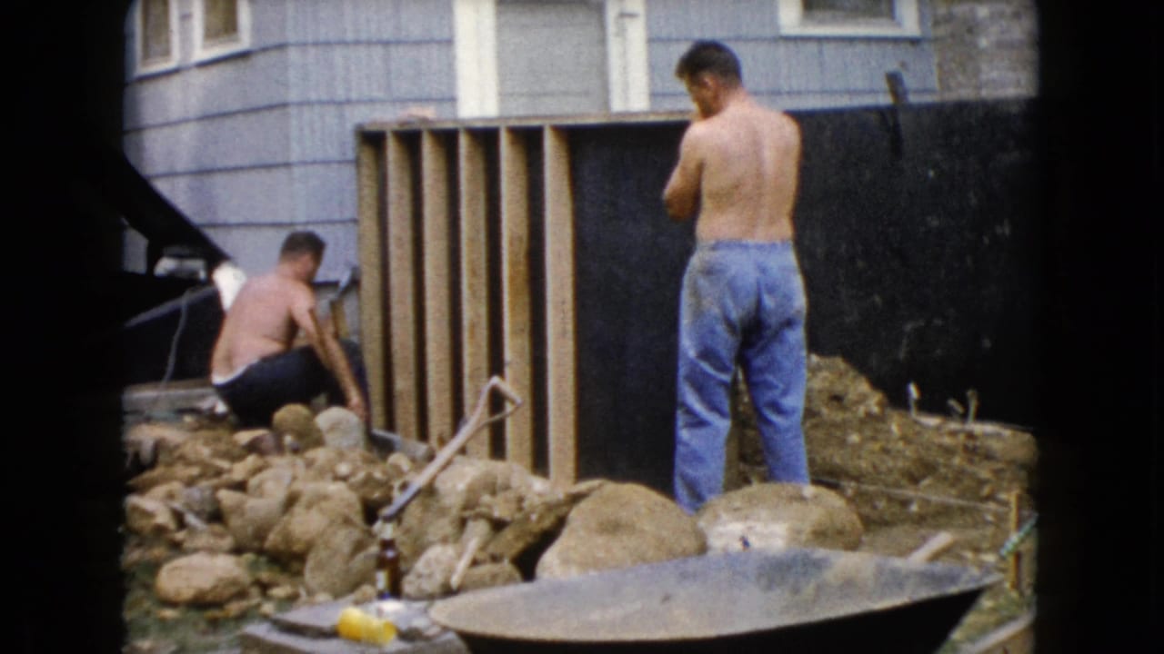 A 1960 Wauconda yard scene shows one man kneeling with a tool at a wooden frame while another stands nearby in blue jeans.