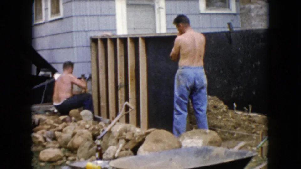 The home-movie frame shows backyard construction activity in Wauconda, with a kneeling man at the wood frame and another man standing beside it.