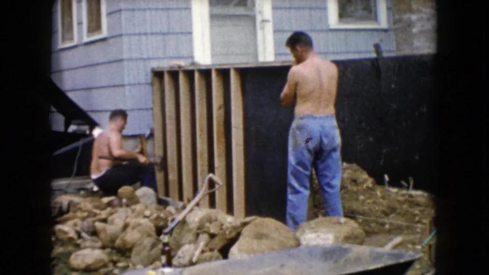 A 1960 Wauconda 8mm frame shows two shirtless men near a partly built wooden structure, with rocks, siding, and yard tools visible.