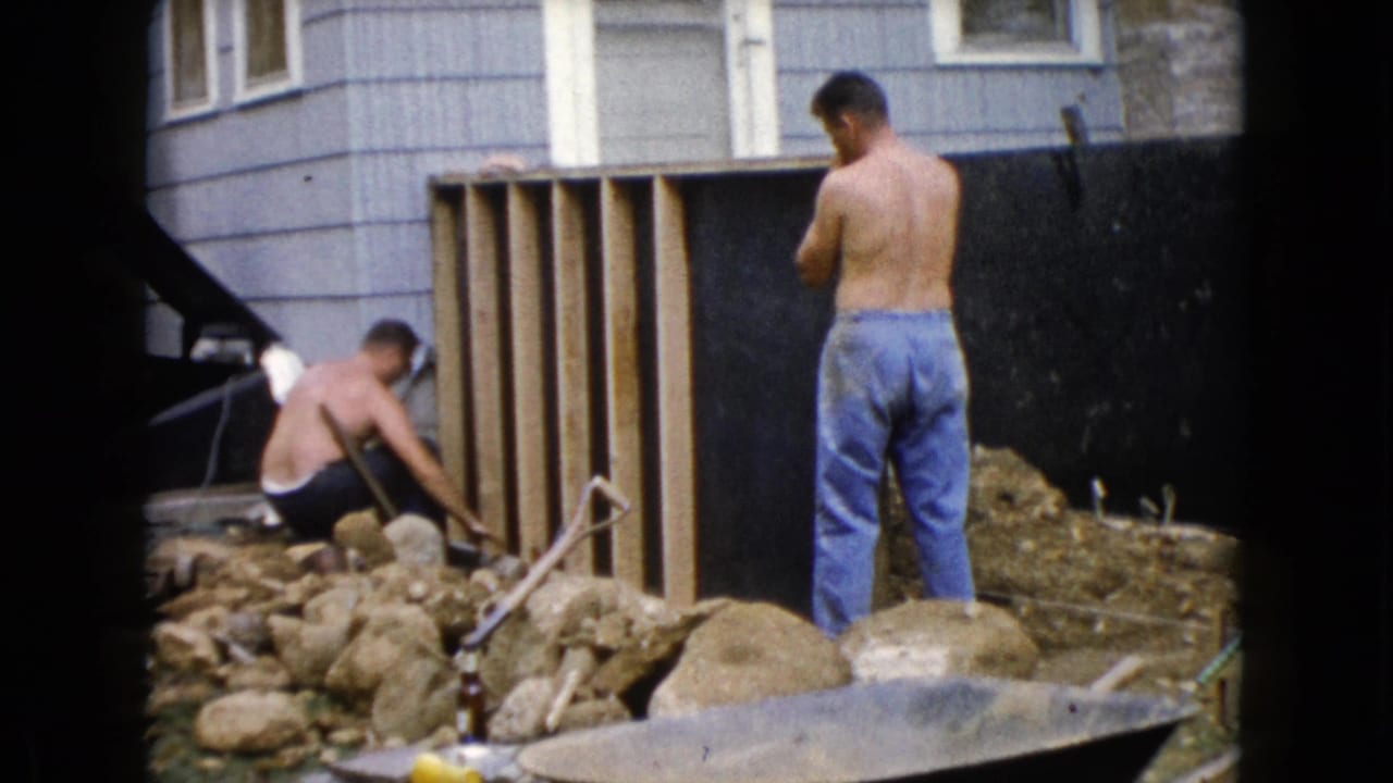 1960 Wauconda, Illinois 8mm frame showing two shirtless men working outside by wood framing, rocks, and a wheelbarrow beside a house.