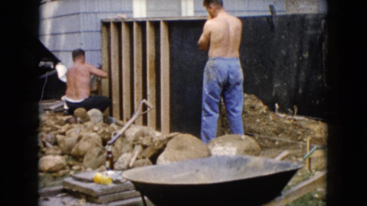 One man kneels at the wood frame while another stands nearby, with rocks and a wheelbarrow in front.