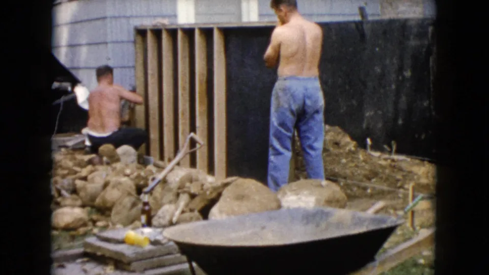 1960 Wauconda, Illinois home-movie frame of one man kneeling at wood framing while another stands near rocks and a wheelbarrow.