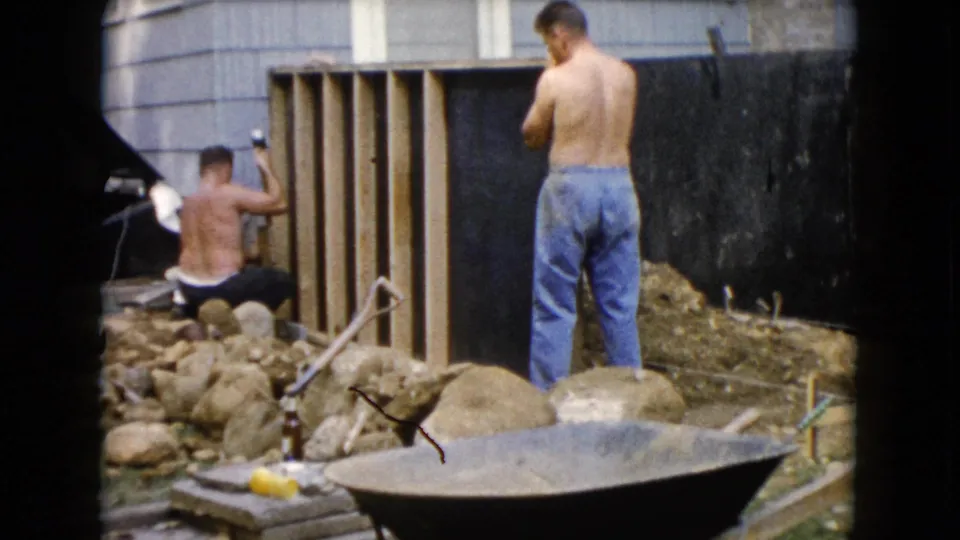 1960 Wauconda, Illinois frame showing a kneeling man lifting a hammer at a wooden frame as another man stands nearby.