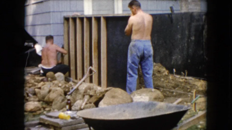 1960 Wauconda, Illinois 8mm frame with two men beside a partially built wooden structure, rocks, and a wheelbarrow.