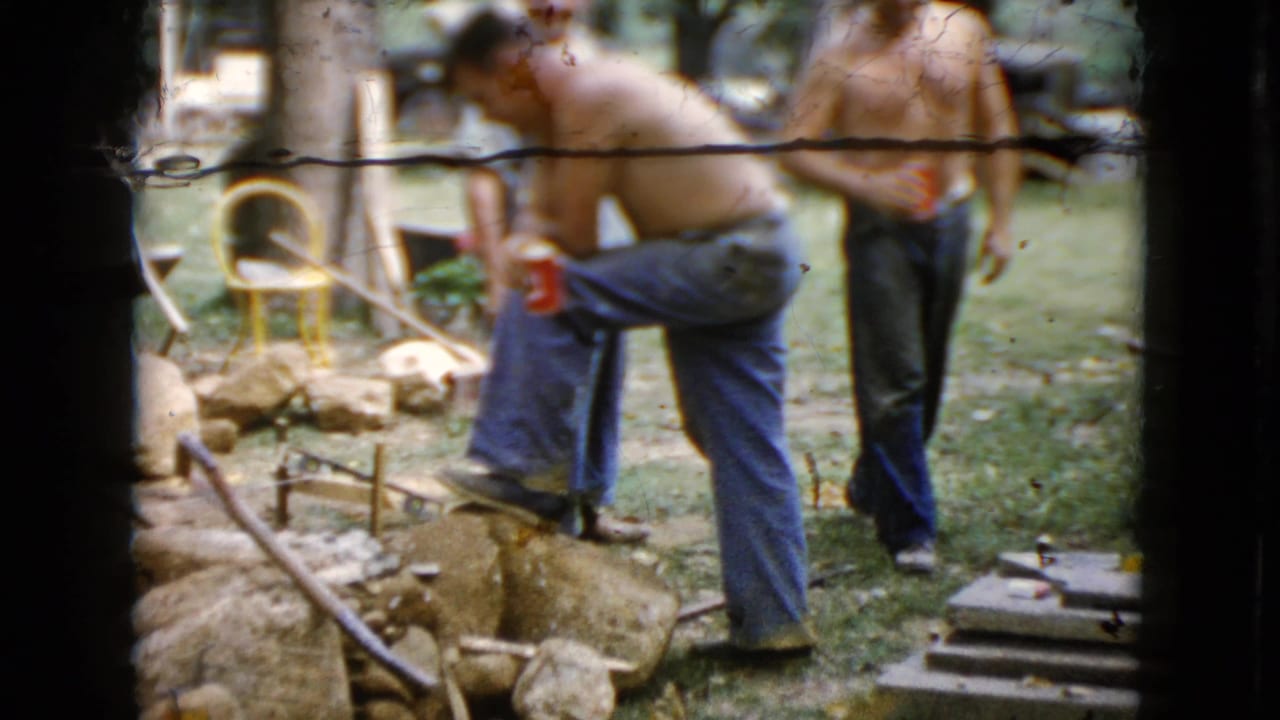 Shirtless men stand and drink near rocks, tools, and a lawn chair in a yard.