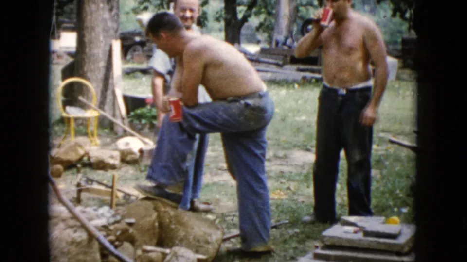 A 1960 backyard work scene in Wauconda shows one man stepping over stones while another drinks from a can beside tools and stacked materials.