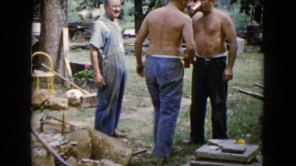 Three adult men gather in a Wauconda yard during a 1960 construction project, with rocks, tools, trees, and stacked slabs visible.
