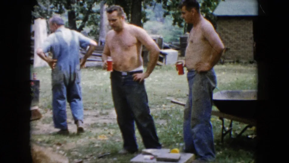 In the 1960 Wauconda home-movie frame, adult men stand near a wheelbarrow, red cups, boards, and yard tools during a work pause.