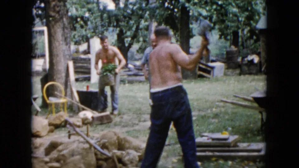 A Wauconda yard work moment shows one shirtless man swinging a sledgehammer as another adult man carries plants near rocks and tools.