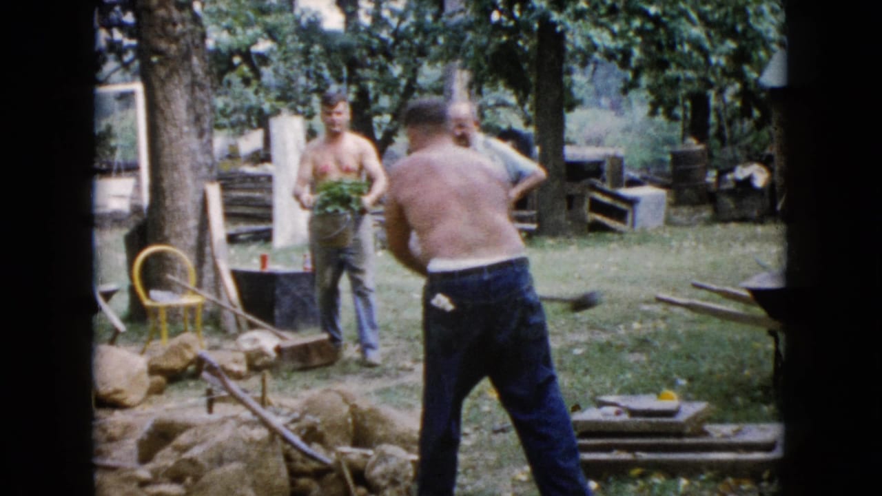 1960 Wauconda, Illinois home-movie frame showing adult men working in a grassy backyard with rocks, tools, and plants.