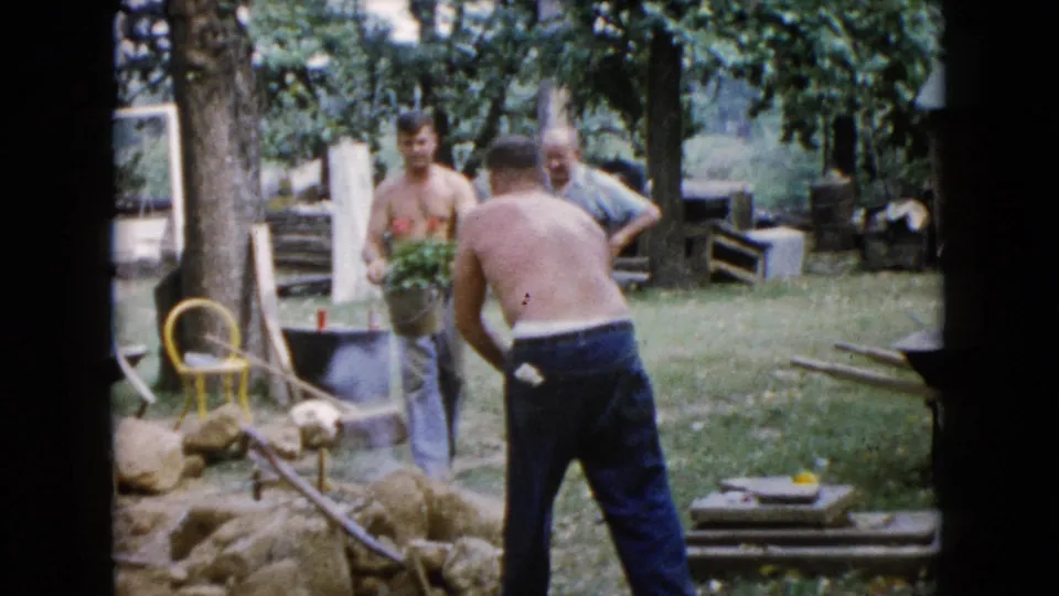 1960 Wauconda backyard scene with men handling a shovel and a bucket of plants near rocks and scattered garden materials.