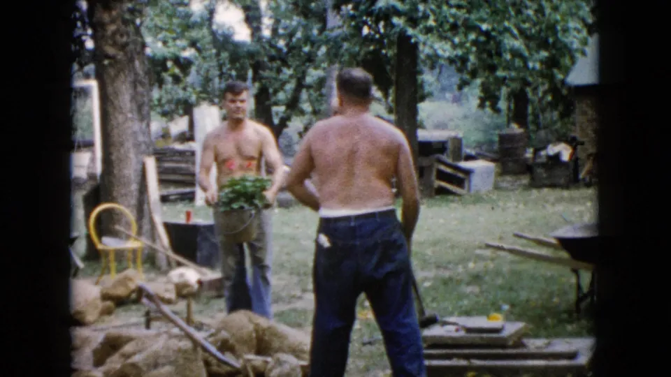 1960 Wauconda home-movie still of men pausing during backyard garden work beside rocks, tools, and a yellow chair.