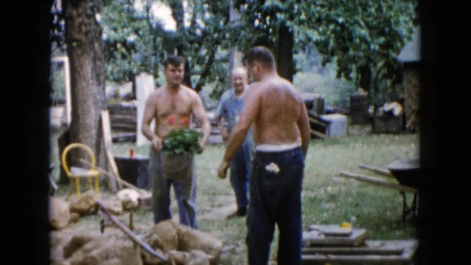 1960 Wauconda, Illinois frame showing three men in a backyard garden area with plants, rocks, tools, and trees.