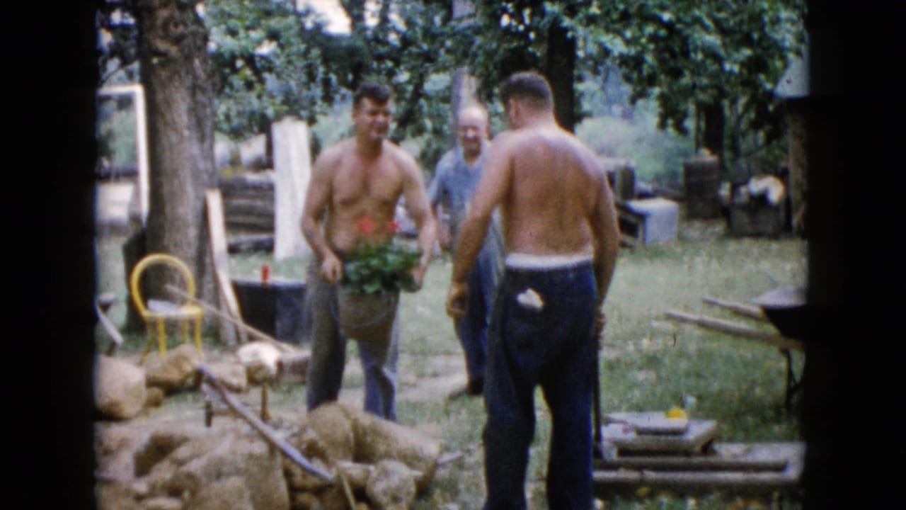 1960 Wauconda 8mm frame showing three adult men in a yard, one shirtless man holding a plant while another stands near rocks and tools.