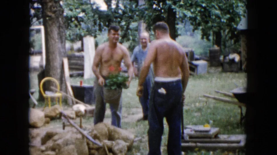 1960 Wauconda 8mm frame showing three adult men in a yard, one shirtless man holding a plant while another stands near rocks and tools.