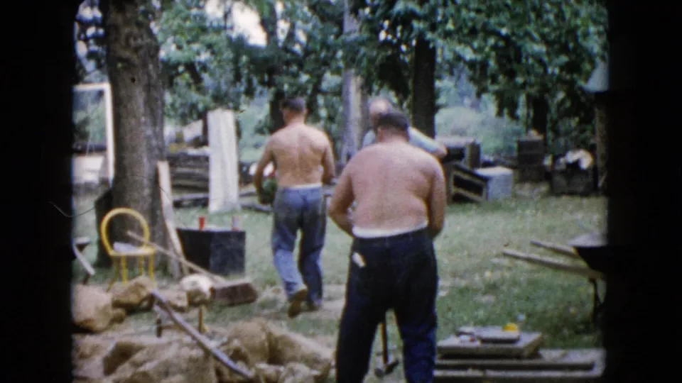 1960 Wauconda 8mm frame showing a shirtless man walking across a grassy yard with another adult man near scattered work materials.