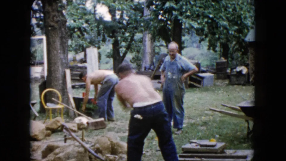 1960 Wauconda backyard frame with shirtless men bending over rocks and plants as a man in a blue shirt observes nearby.