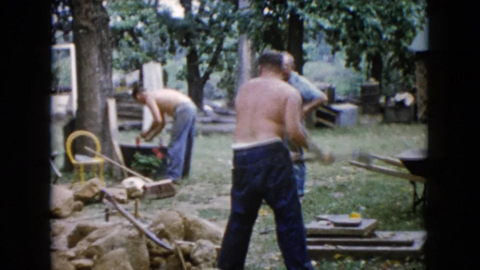 1960 Wauconda home-movie frame showing men outdoors with rocks, tools, a wheelbarrow, and a yellow chair in a shaded yard.