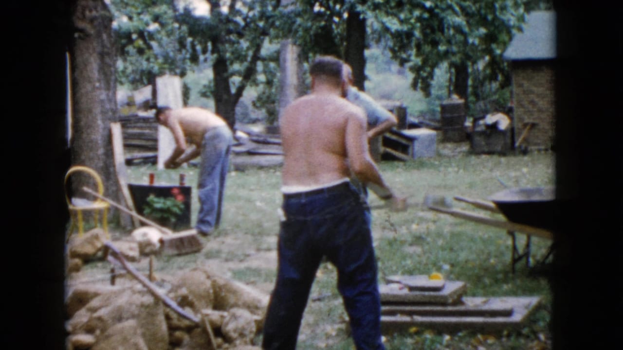 1960 Wauconda backyard scene with a shirtless man near stones and another man bent over a black container beside tools and a wheelbarrow.