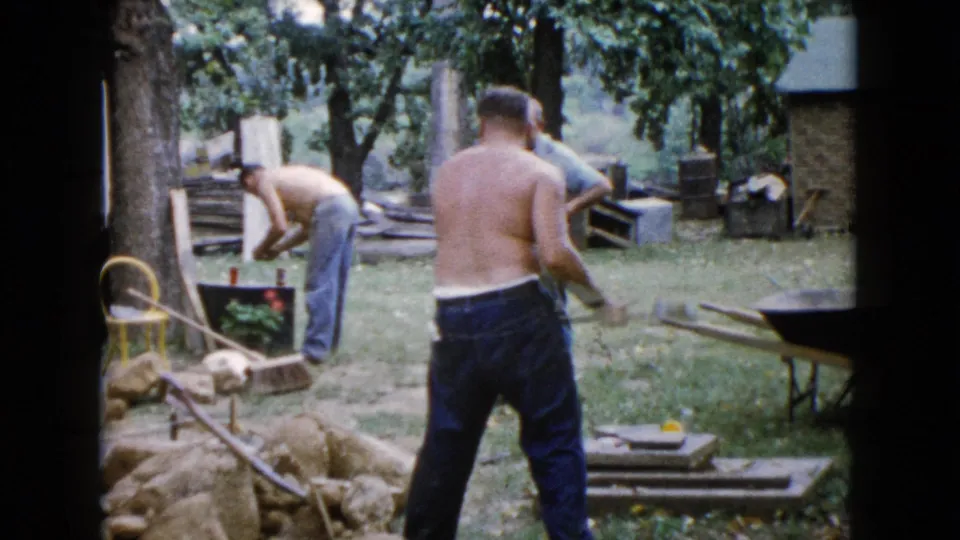 1960 Wauconda backyard scene with a shirtless man near stones and another man bent over a black container beside tools and a wheelbarrow.