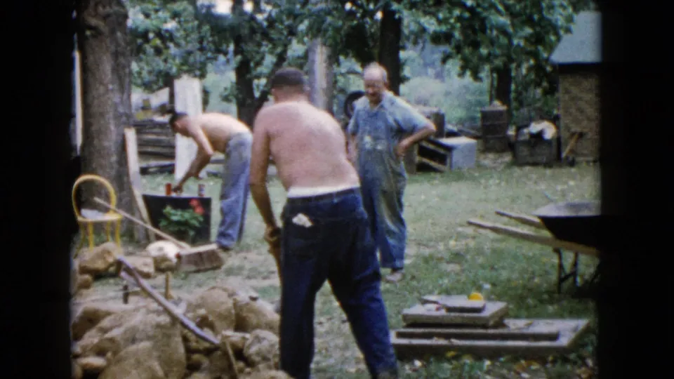 1960 Wauconda 8mm frame showing three adult men in a backyard with rocks, tools, a wheelbarrow, and trees around them.