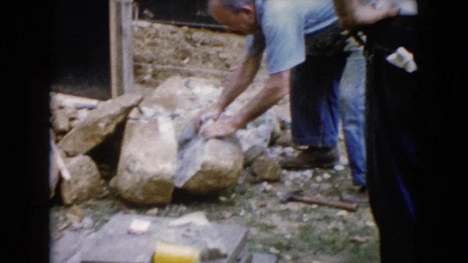 Close 1960 Wauconda home-movie frame of a man leaning over a large stone in a yard, with another figure partly visible at the edge.