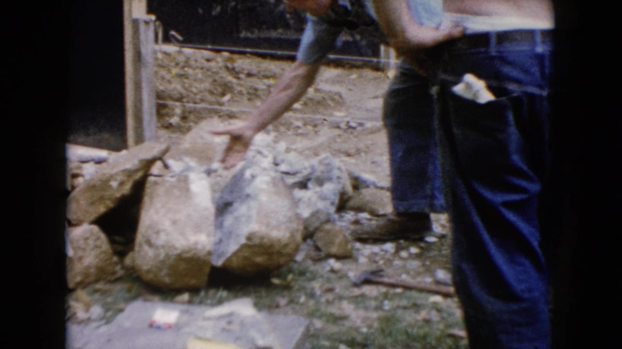 Man in jeans bends near a pile of large stones at an outdoor work area.