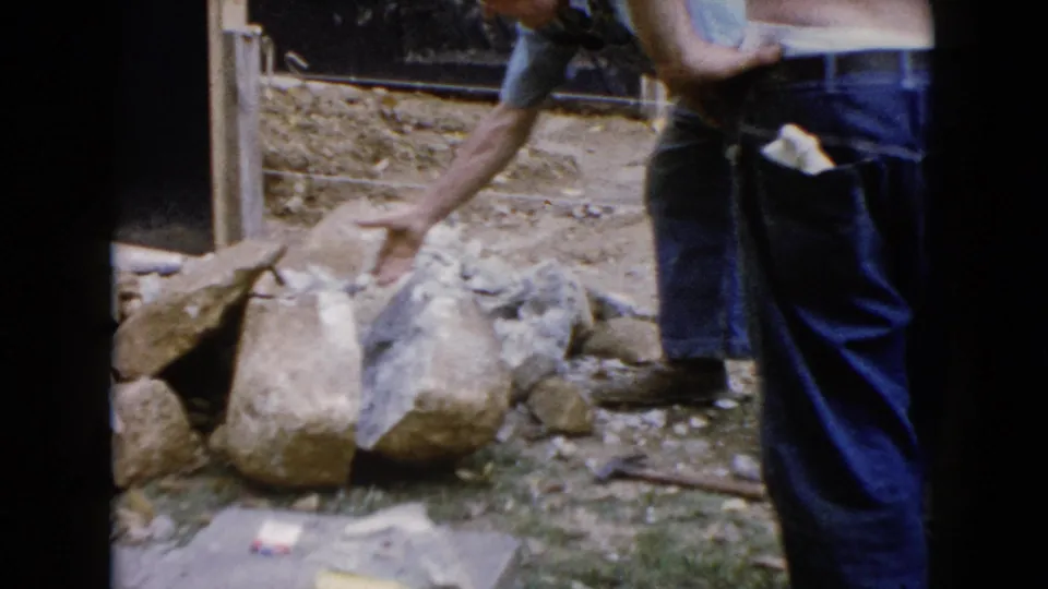 In 1960 Wauconda, Illinois footage, a man bends beside large rocks and loose dirt at a small outdoor construction site.