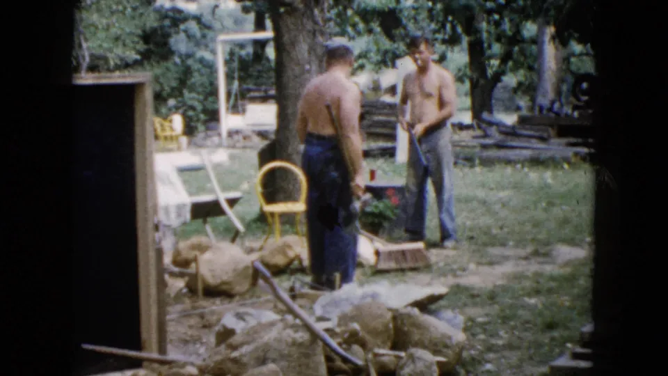 1960 Wauconda 8mm frame showing two shirtless adult men working among rocks and tools in a grassy backyard.