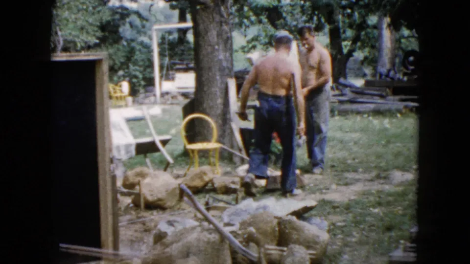 Two adult men in a 1960 Wauconda backyard stand beside rocks, tools, trees, and a yellow chair during outdoor work.