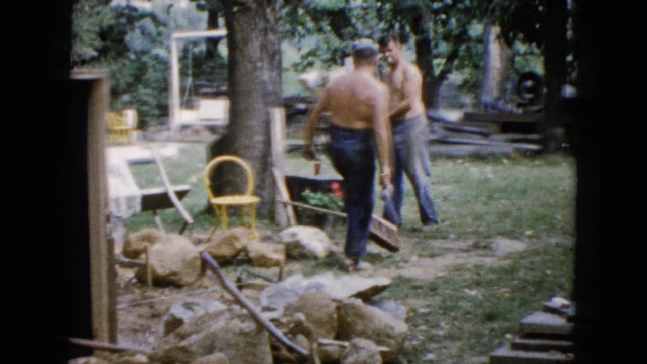 In 1960 Wauconda, Illinois home-movie footage, two shirtless men work in a backyard near rocks, tools, lawn furniture, and trees.