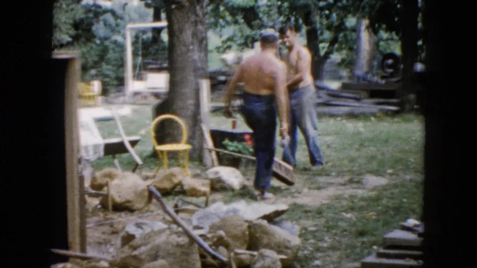 In 1960 Wauconda, Illinois home-movie footage, two shirtless men work in a backyard near rocks, tools, lawn furniture, and trees.