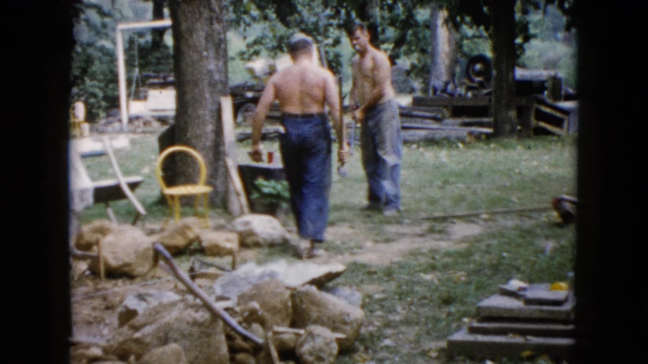 Two adult men appear to talk or coordinate while working in a 1960 Wauconda backyard with rocks, tools, trees, and lawn furniture nearby.