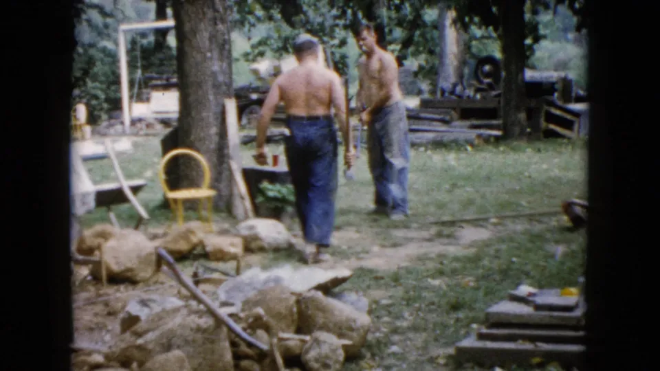 Two adult men appear to talk or coordinate while working in a 1960 Wauconda backyard with rocks, tools, trees, and lawn furniture nearby.