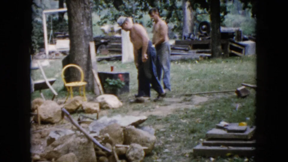 A shirtless man bends near the ground as another stands behind him during backyard work in 1960 Wauconda, Illinois home-movie footage.