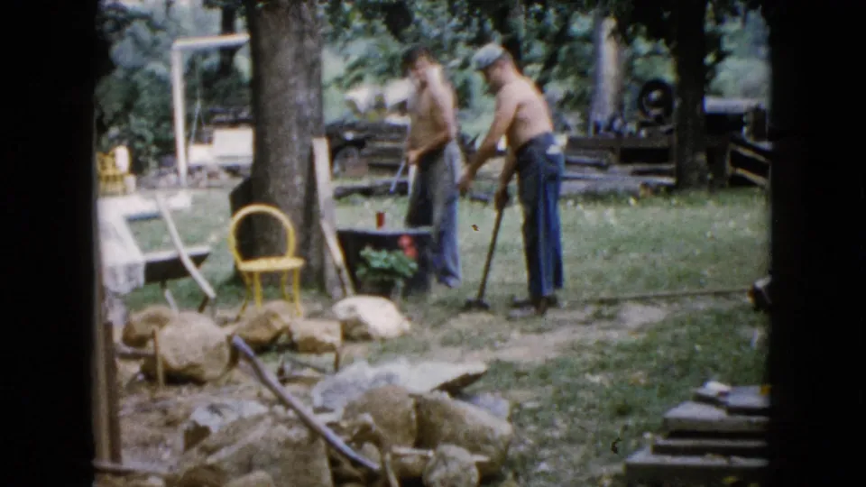 Two men handle long tools in a Wauconda backyard, with rocks, trees, a yellow chair, and lawn equipment visible in the 1960 8mm frame.